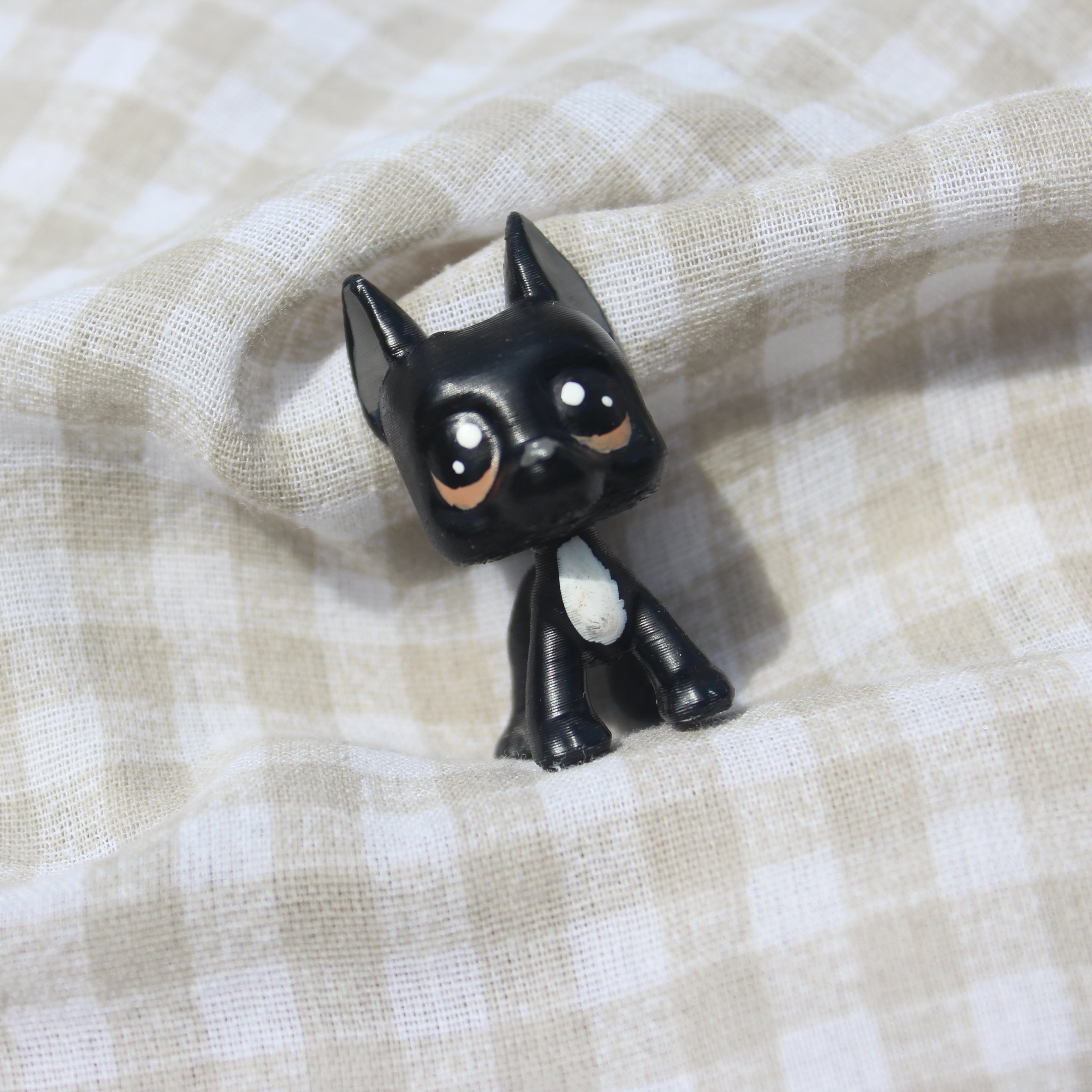 A small black great dane dog figurine with large white and brown eyes and a white chest patch, posed on a beige and white checkered fabric background.