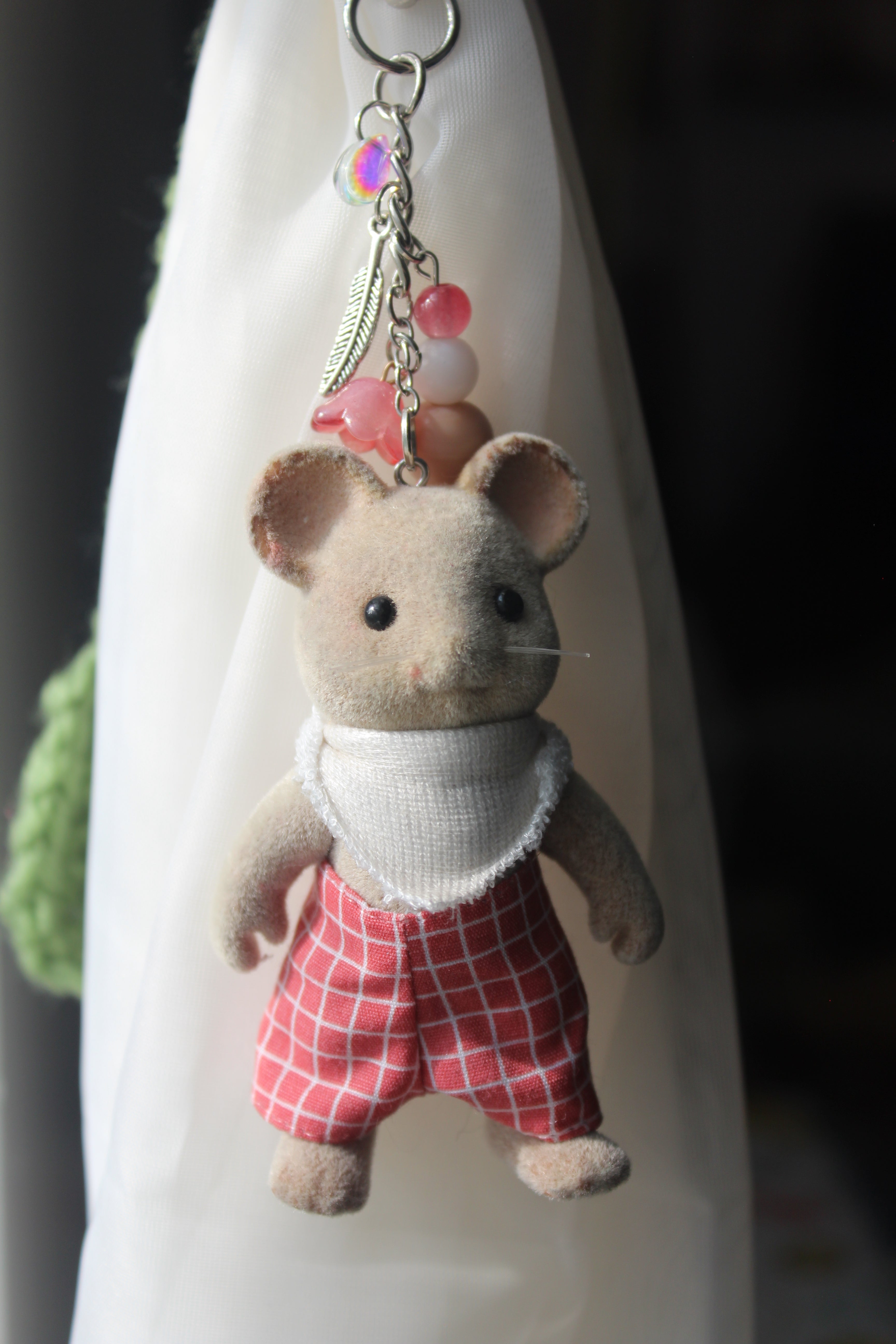 A small plush mouse keychain or bag charm with a beige body, wearing a white top and red-and-white checkered pants, attached to a silver metal ring with pink beads.