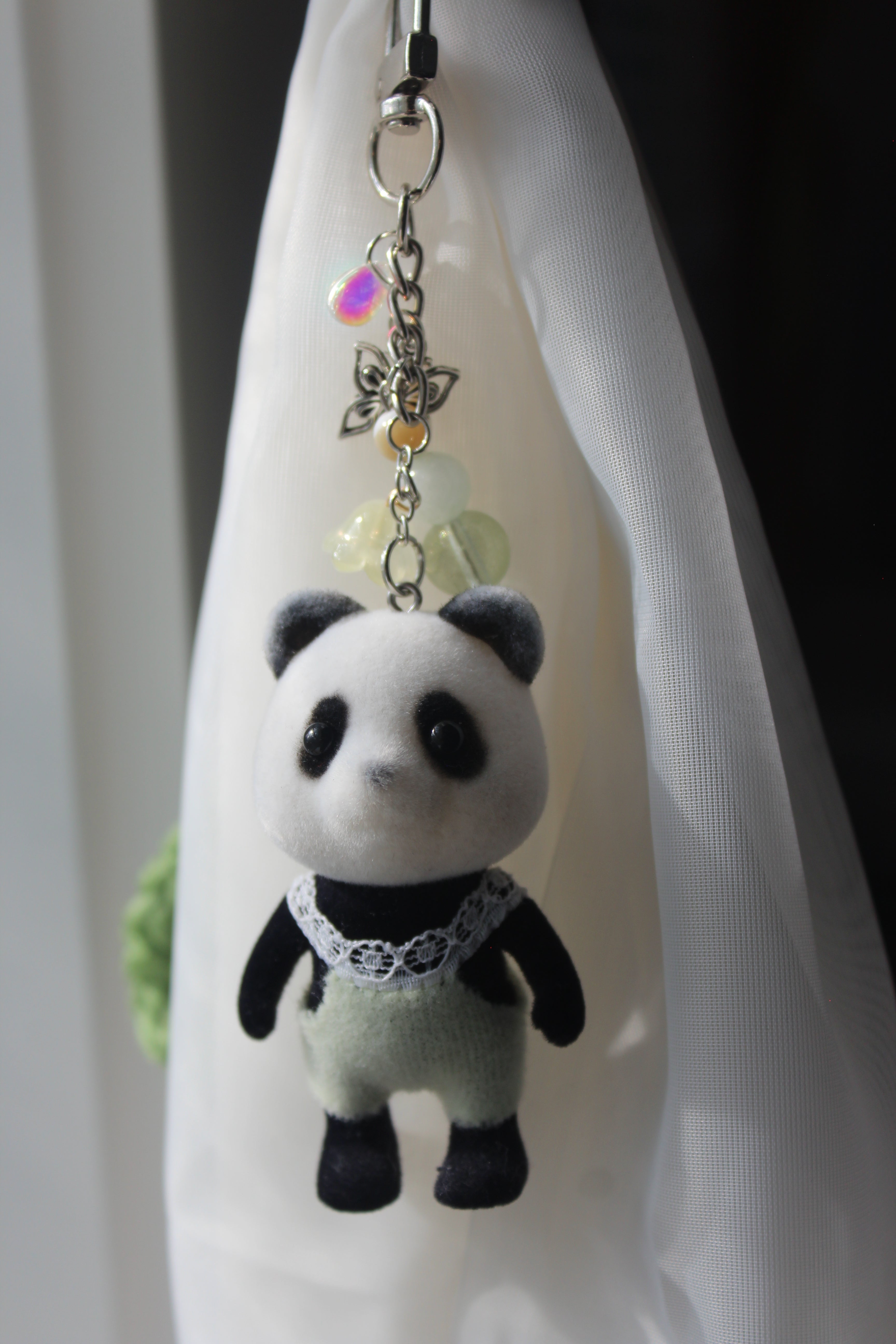 A small plush panda keychain or bag charm with black and white fur, yellow and lace outfit, and a silver metal clasp with iridescent beads, resting on a white fabric surface.