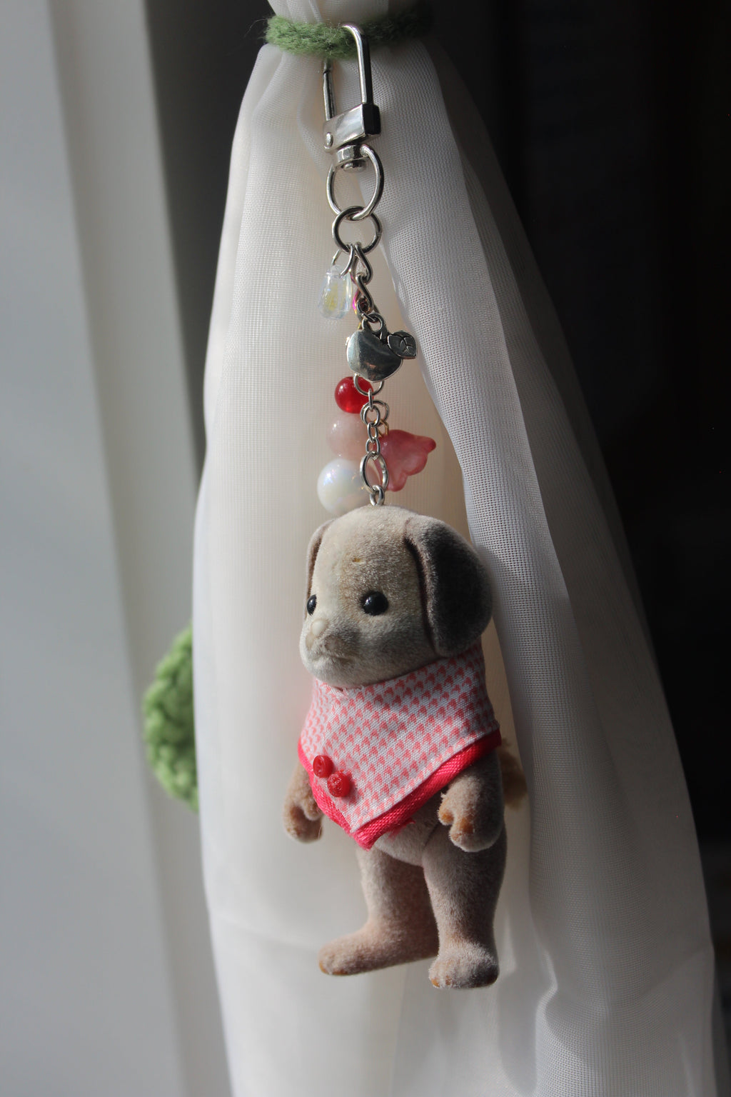 A small plush dog keychain accessory with a red and white gingham dress, lying on a white fabric surface near a window.