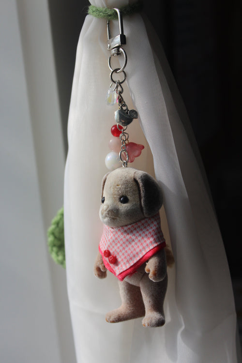 A small plush dog keychain accessory with a red and white gingham dress, lying on a white fabric surface near a window.