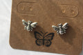 A pair of silver butterfly stud earrings displayed on a brown kraft paper backing card with a black butterfly graphic, set against a white textured background.